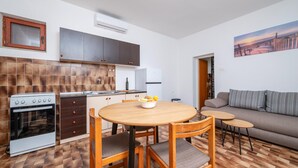A compact kitchen with a stove, oven, and modern cabinetry. There is a dining area with a wooden table and chairs, and a small round table with a chair. A sofa and a painting are visible in the living area. - Awesome apartment in Fazana (Fazana)