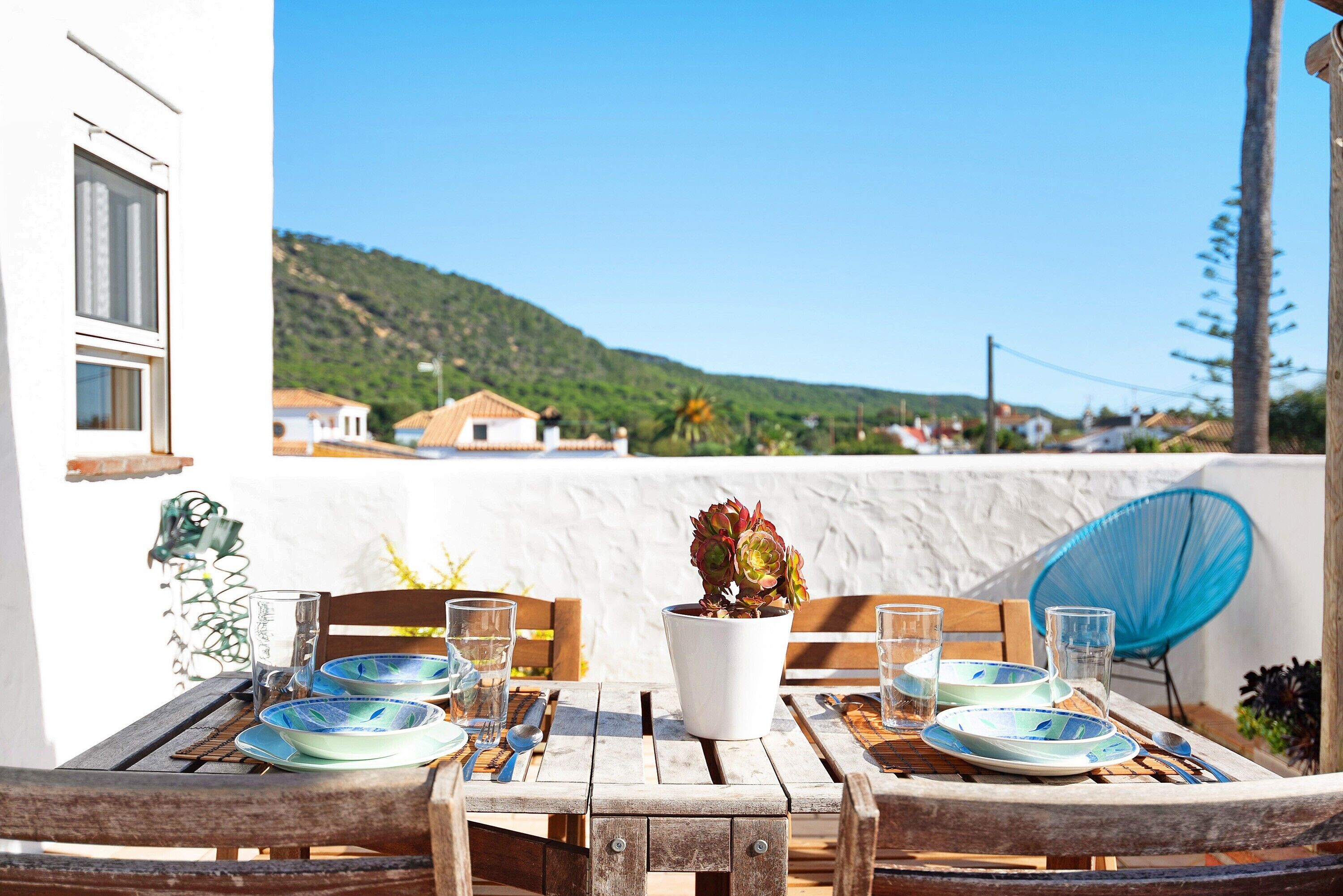 A wooden table with plates and glasses, a potted plant, and a view of a hillside.