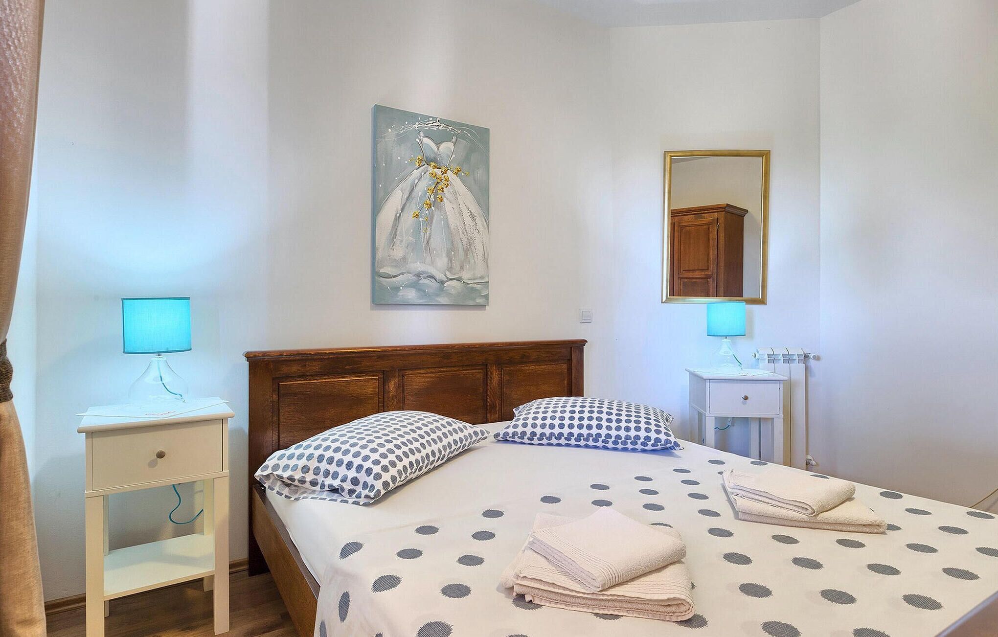 A bedroom with a wooden headboard, a bed with polka dot bedding, two bedside tables with lamps, a wall-mounted mirror, and a painting above the bed.