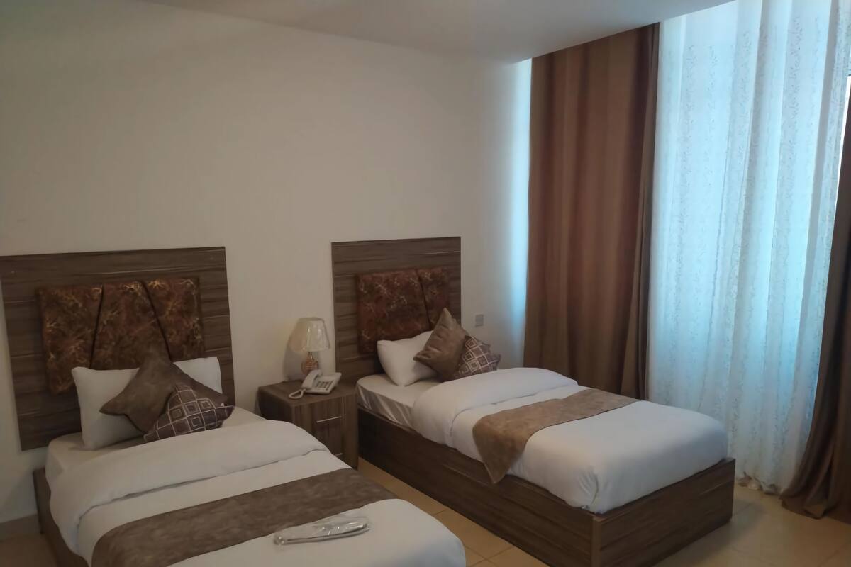 Ein Hotelzimmer mit zwei Betten, einem Nachttisch mit Lampe und Telefon sowie einem Fenster mit Vorhängen.