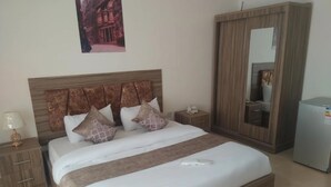 A hotel room with a large bed, a wooden headboard, a wardrobe, a bedside table with a lamp, and a small refrigerator. - Rayan Hotel Suites (Amman)