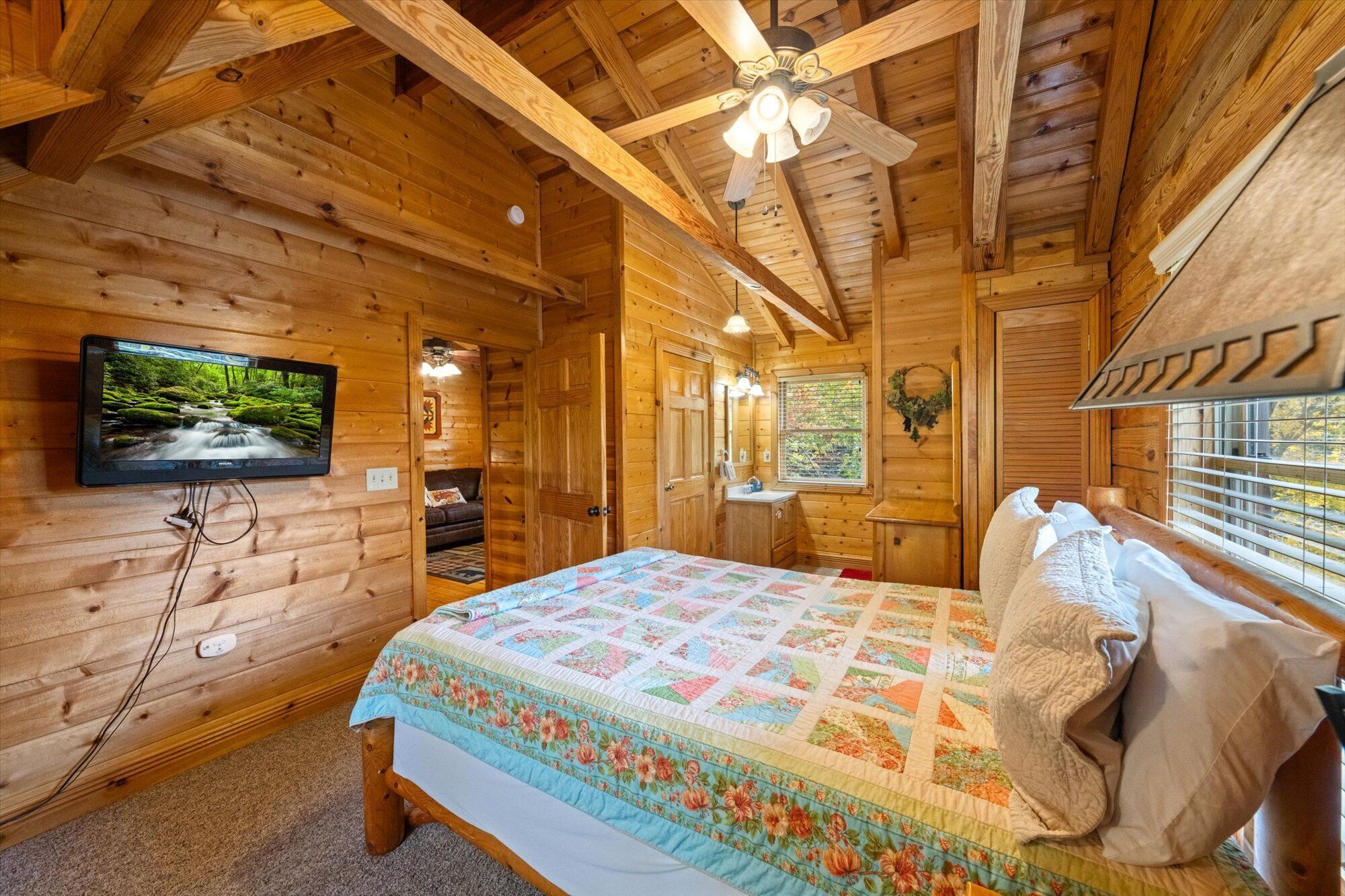 A wooden cabin bedroom with a bed, a television, a ceiling fan, and a window with blinds.