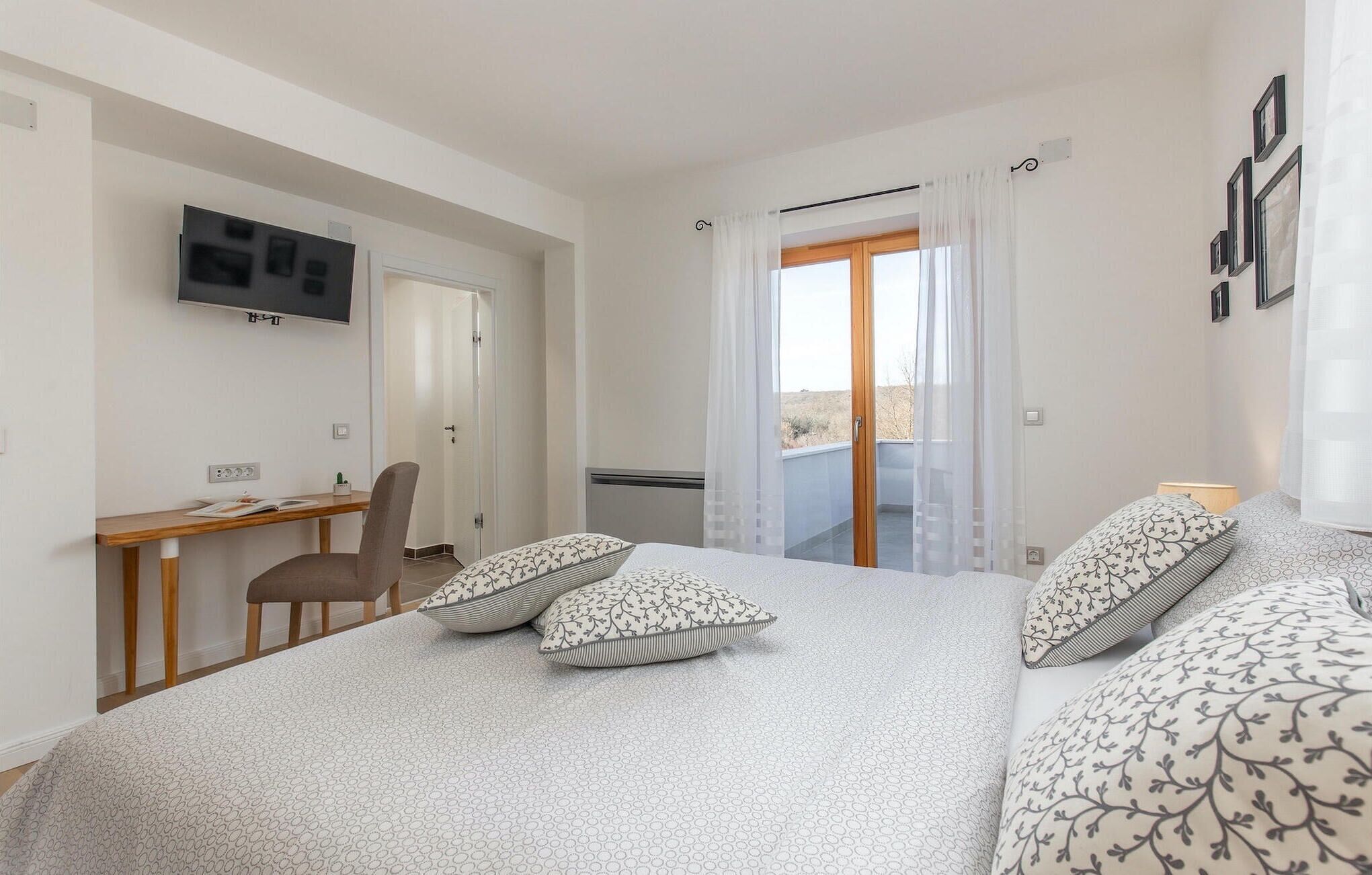 A bedroom with a bed, a desk, a chair, a TV, and a balcony with a view.