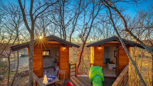 Deux cabanes en bois reliées par une terrasse en bois, le tout entouré d’arbres.