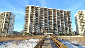 A high-rise residential building with multiple balconies, a wooden boardwalk leading to the entrance, and a clear sky. - Snowbird Availability!  Beachfront 3BR!!  Inquire for best Rates! (Orange Beach)