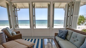 Een kamer aan het strand met rieten meubels, een glazen tafel en uitzicht op de oceaan door geopende luiken.