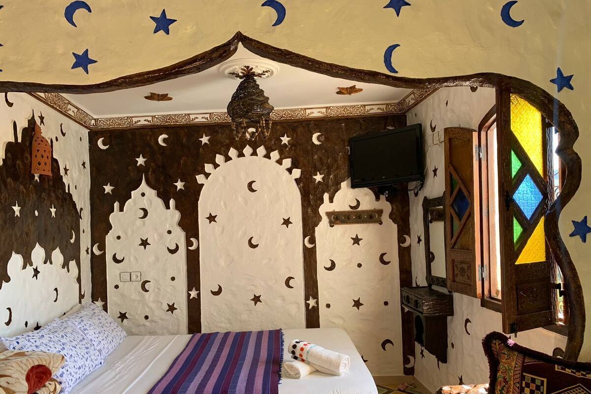 Ein Schlafzimmer mit einem Bett, einem Nachttisch, einem Fernseher und einem Fenster mit geometrischem Muster.