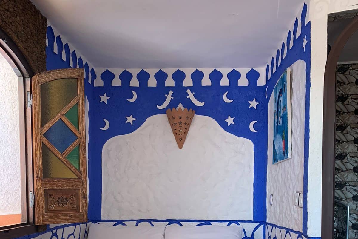 Ein Bett mit weißem Kopfteil und blauen Dekorationselementen, eine Holztür mit geometrischem Muster und eine Steinsturz.