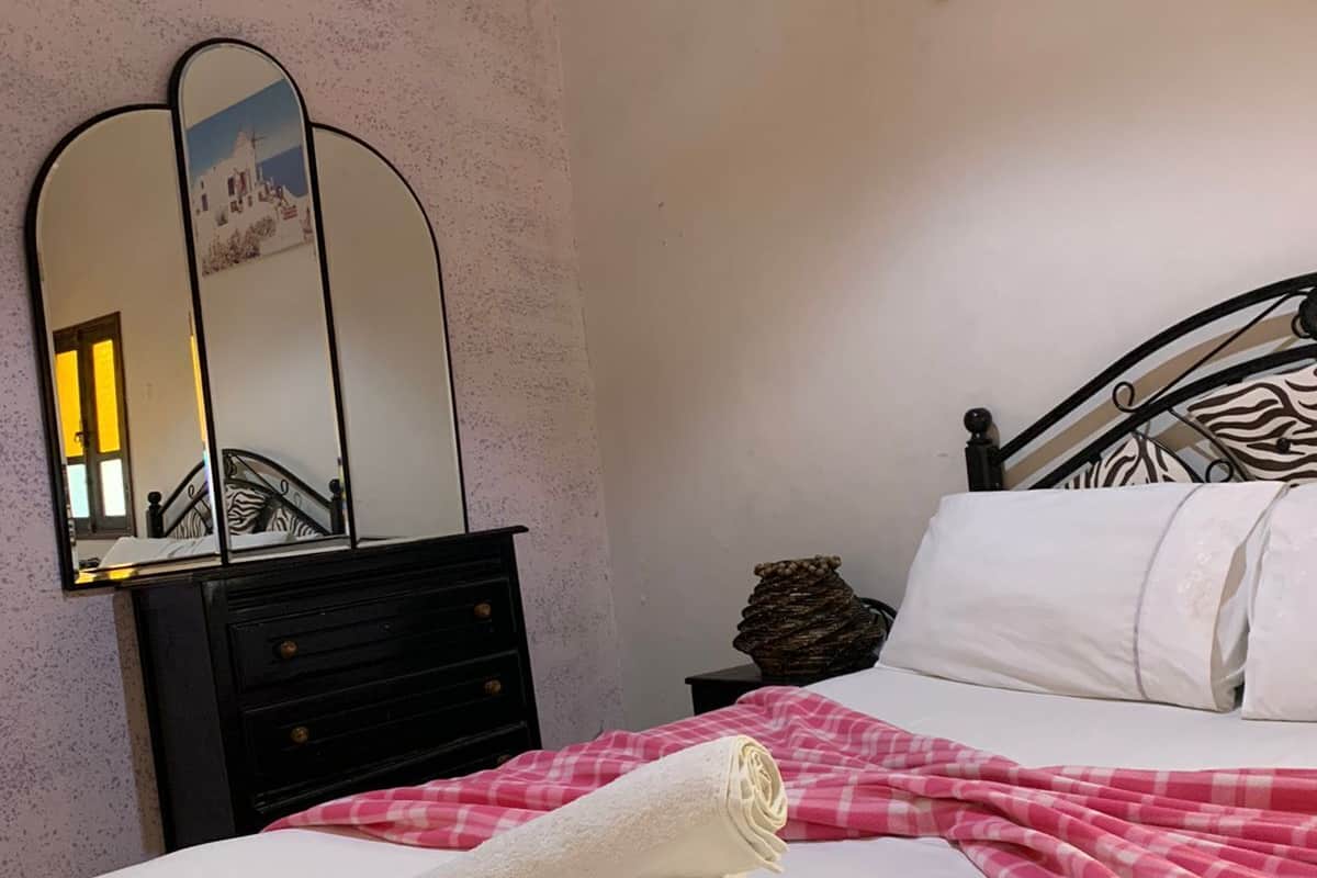 Ein ordentlich bezogenes Bett mit weißen Leinen und einer pink karierten Decke, eine schwarze Kommode mit Spiegel und ein Gemälde einer Küstenlandschaft an der Wand.
