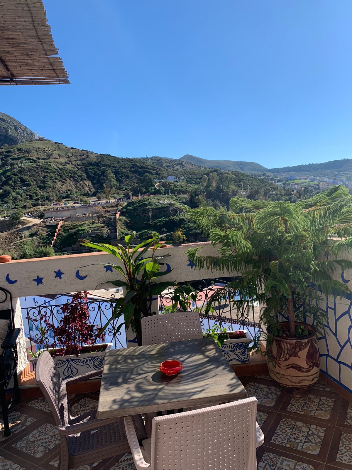 A terrace with a table and chairs, potted plants, and a view of a valley with houses and greenery.