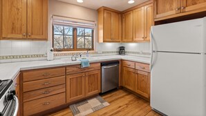 Una cocina con armarios de madera, un refrigerador blanco, un lavavajillas y una ventana con vista a árboles.