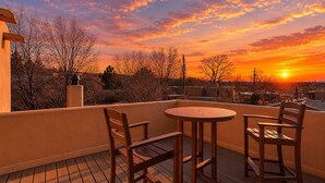 Una terraza de madera con una mesa redonda y dos sillas, con vistas a una zona residencial al atardecer.