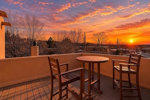 A wooden deck with a round table and two chairs, overlooking a residential area during sunset.