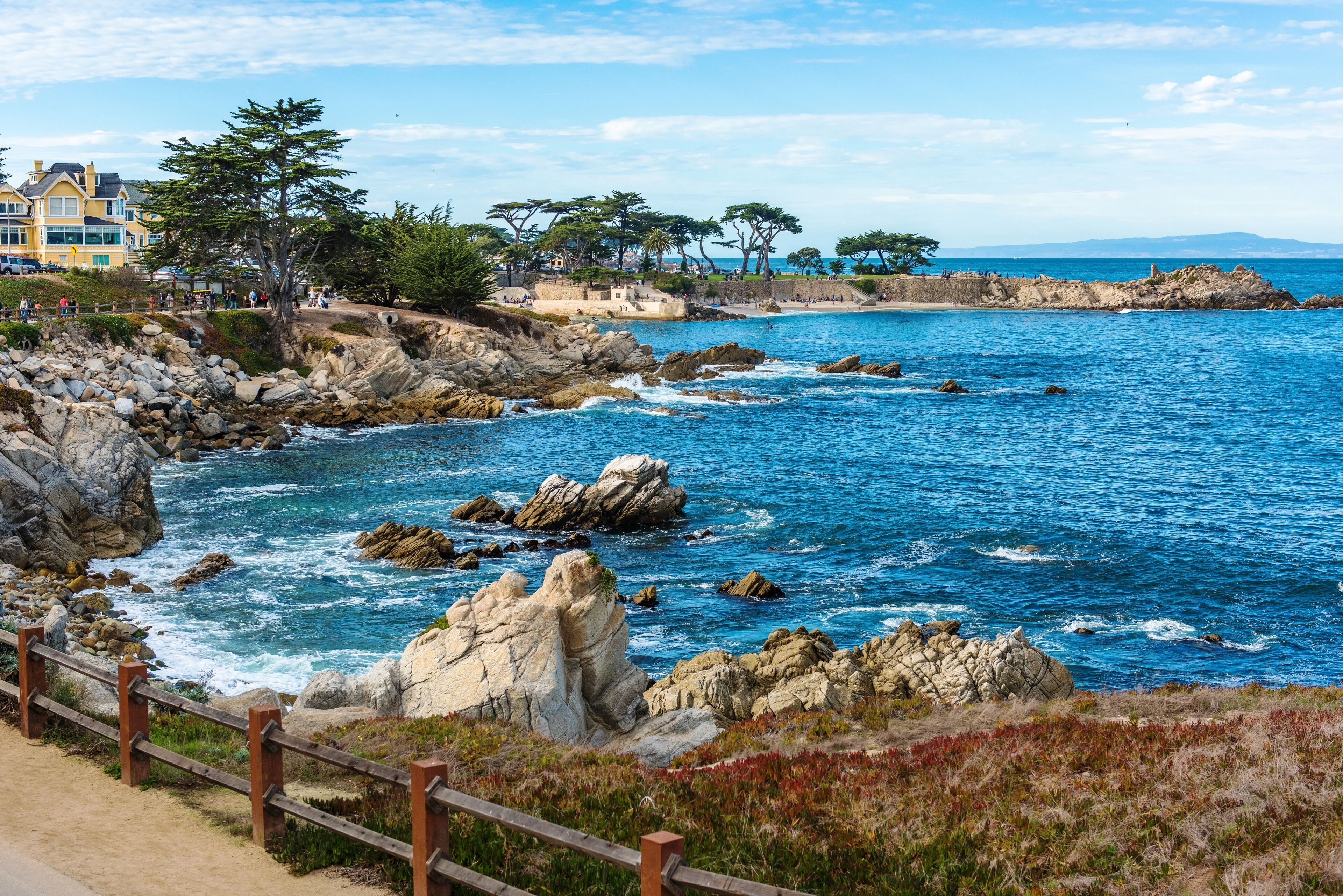 A coastal scene with rocky terrain, a wooden fence, and a clear blue sea.