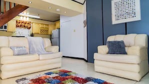 A living room with two cream-colored sofas, a colorful rug, a kitchen in the background, and a blue wall with a framed picture. -  Sweet Kensville (Kingston)