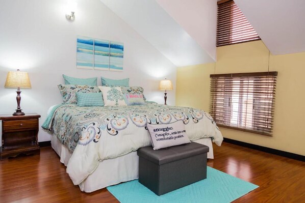 A bedroom with a bed, two bedside lamps, a window with blinds, a painting on the wall, and a ceiling with a sloped design. -  Sweet Kensville (Kingston)
