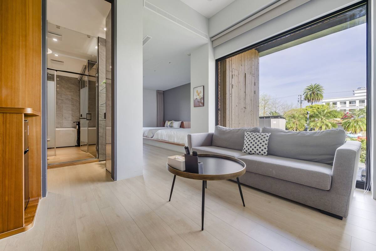 A modern living room with a grey sofa, a round coffee table, and a large window offering a view of the outdoors.