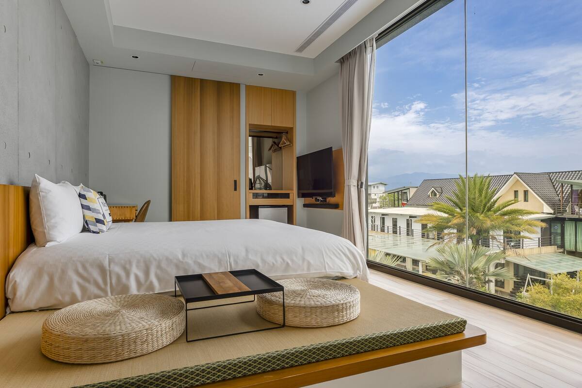 A modern bedroom with a large bed, a TV, and a view of buildings and palm trees outside.