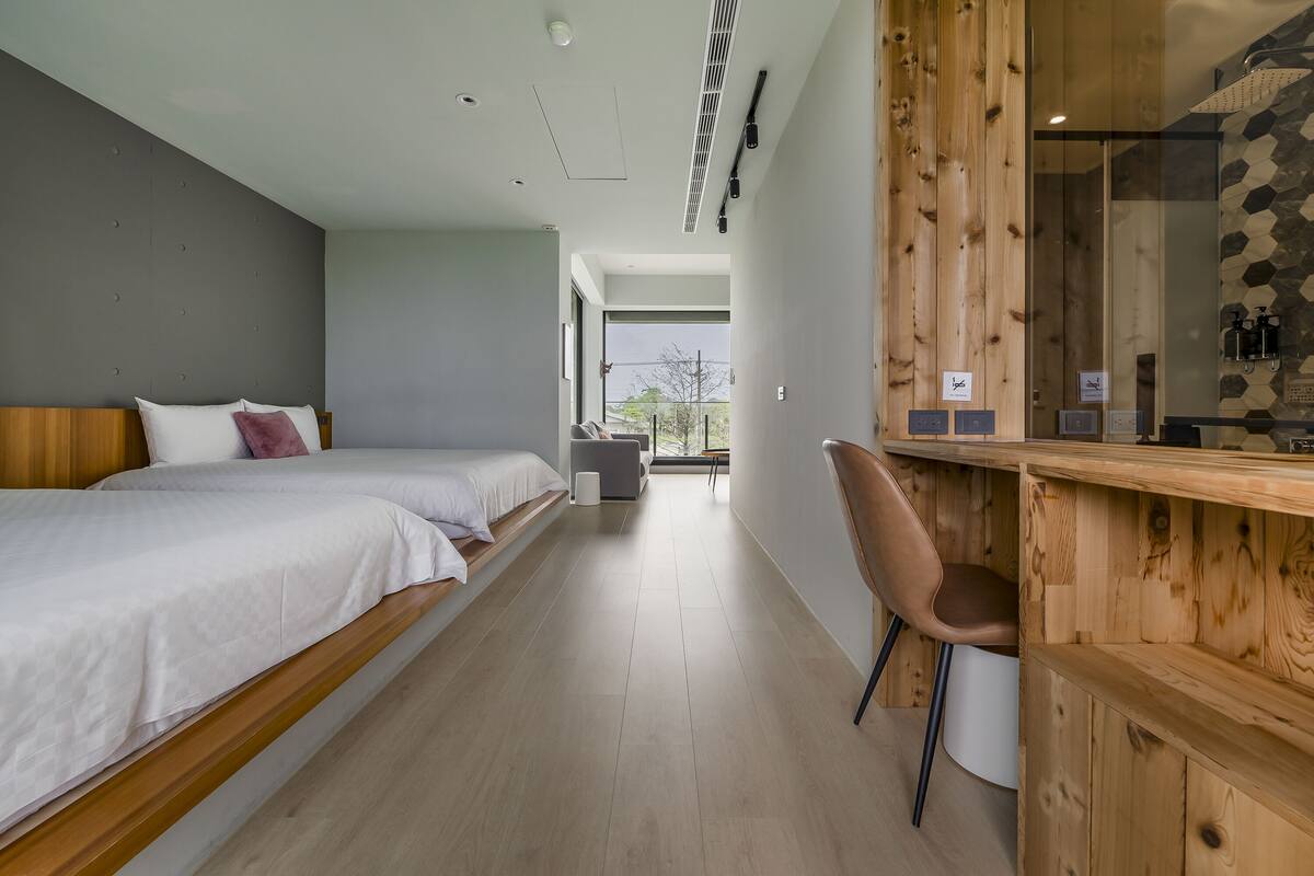 A modern hotel room with two beds, a wooden desk, and a view of the outdoors.