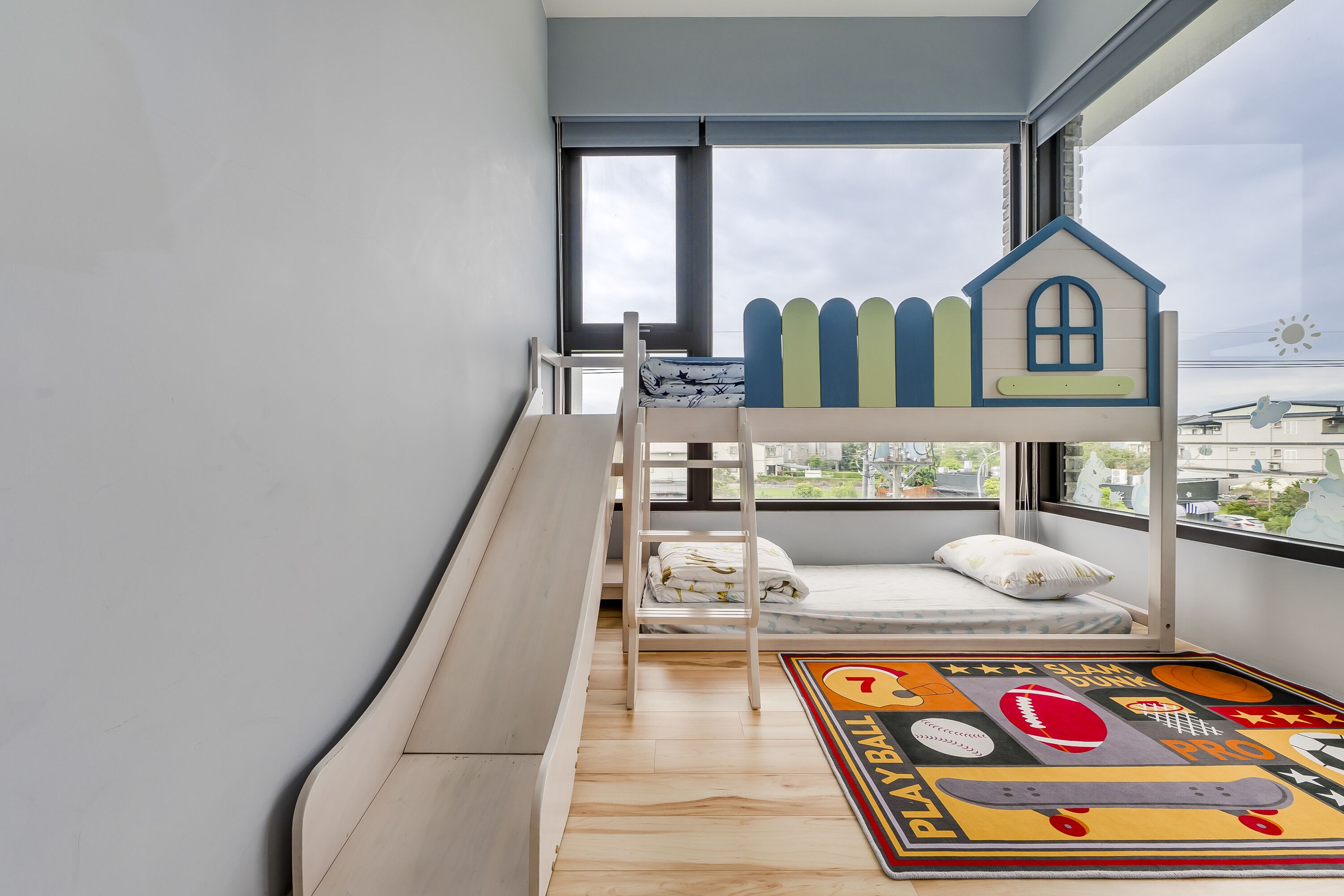 A bunk bed with a small house structure on top, a window with a view of buildings, and a colorful play mat on the floor.