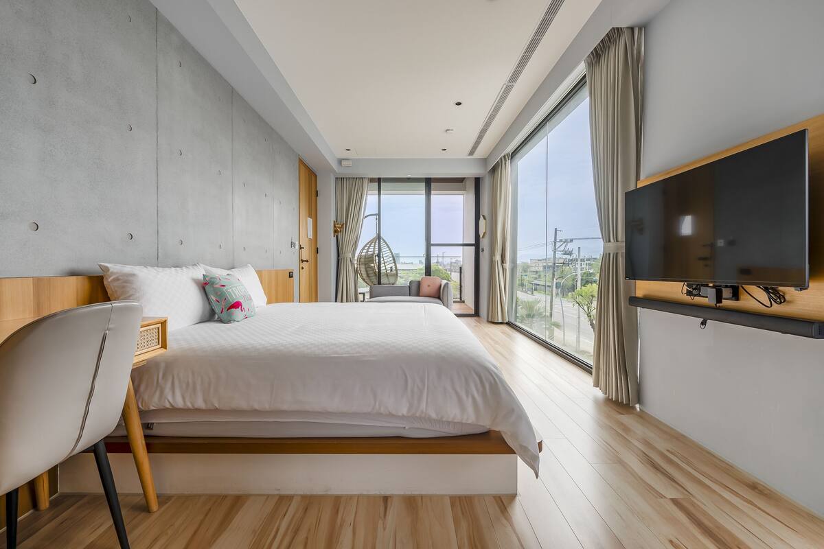 A modern bedroom with a large bed, a TV mounted on the wall, and a view of the city through large windows.