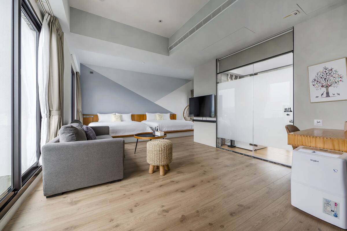 A modern hotel room with a sofa, a small ottoman, a bed, a television, and a refrigerator.