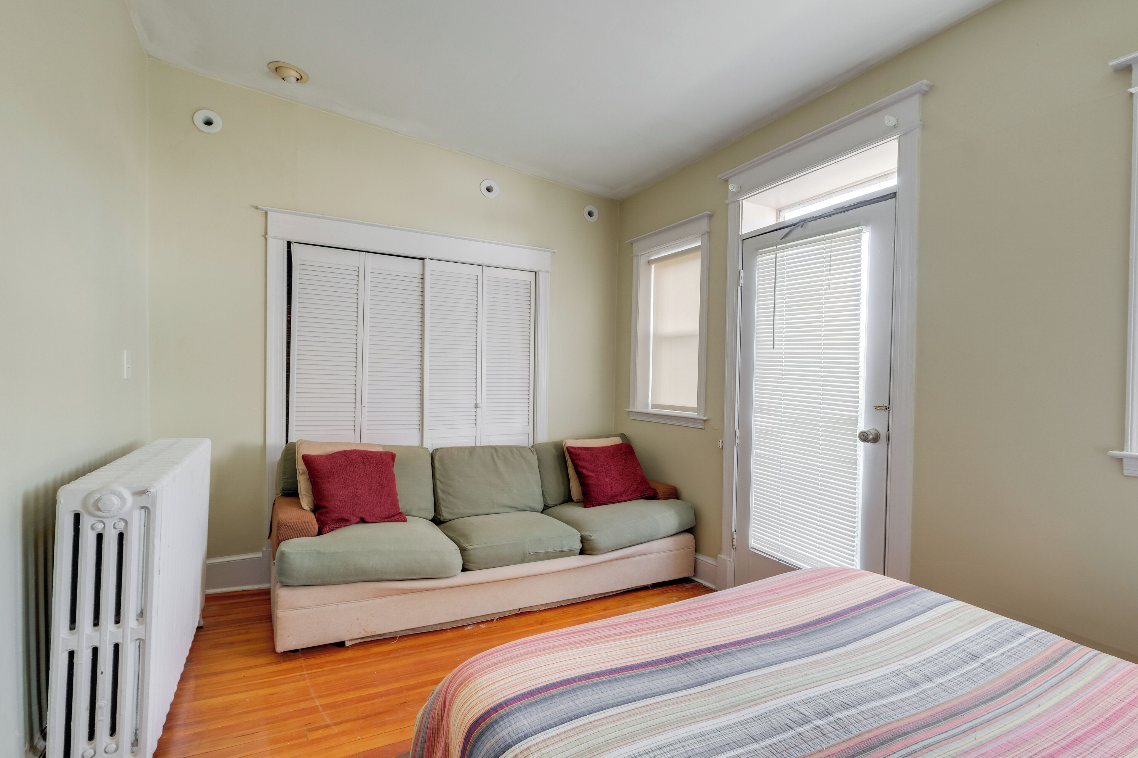 A bedroom with a bed, a radiator, a sofa, and a window with blinds.