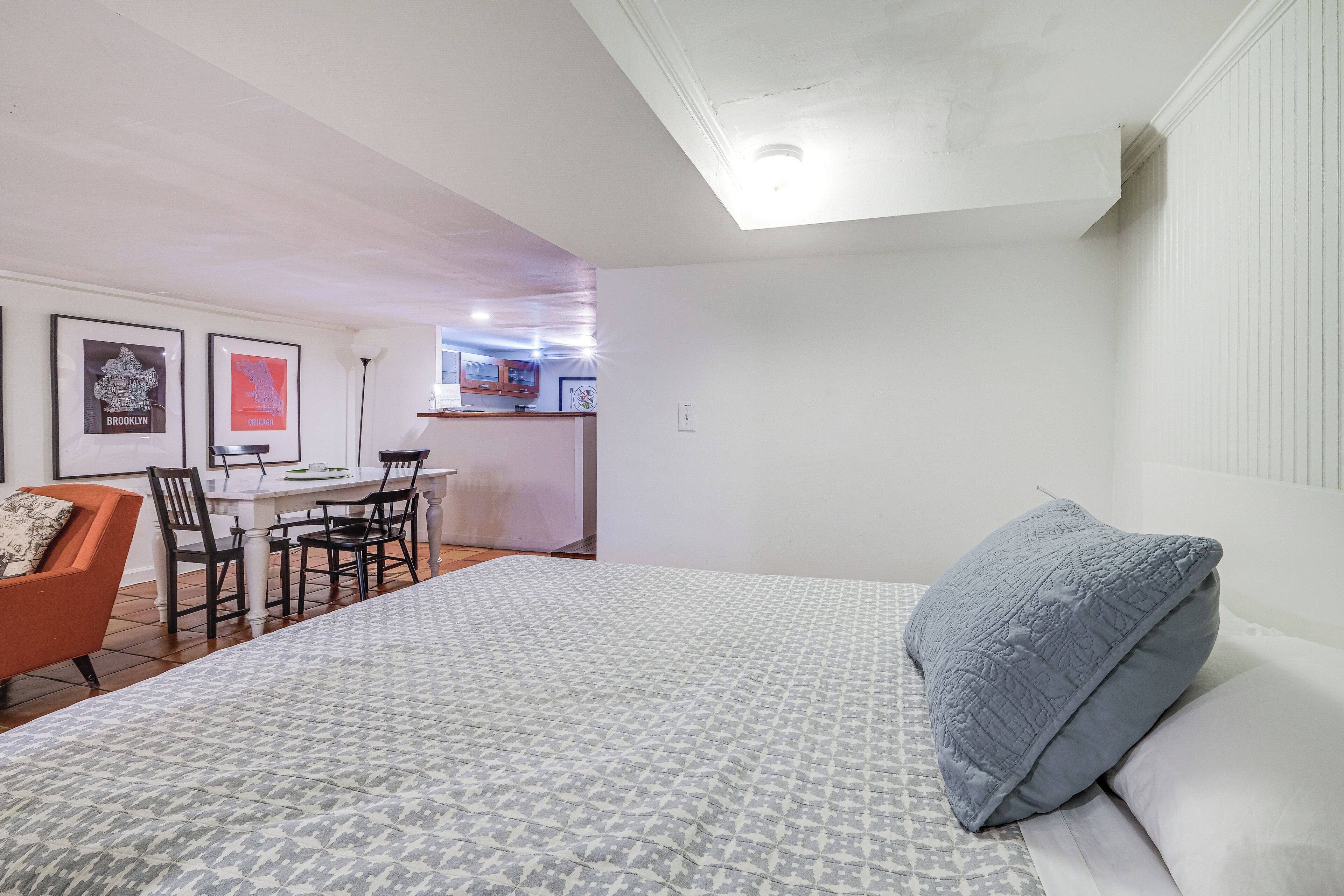 Un dormitorio con cama, un tapete gris con diseño, almohada gris, zona de comedor con mesa y sillas, y cuadros en la pared.