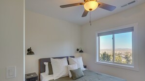 A bedroom with a bed, a ceiling fan, a window with a city view, and bedside lamps.