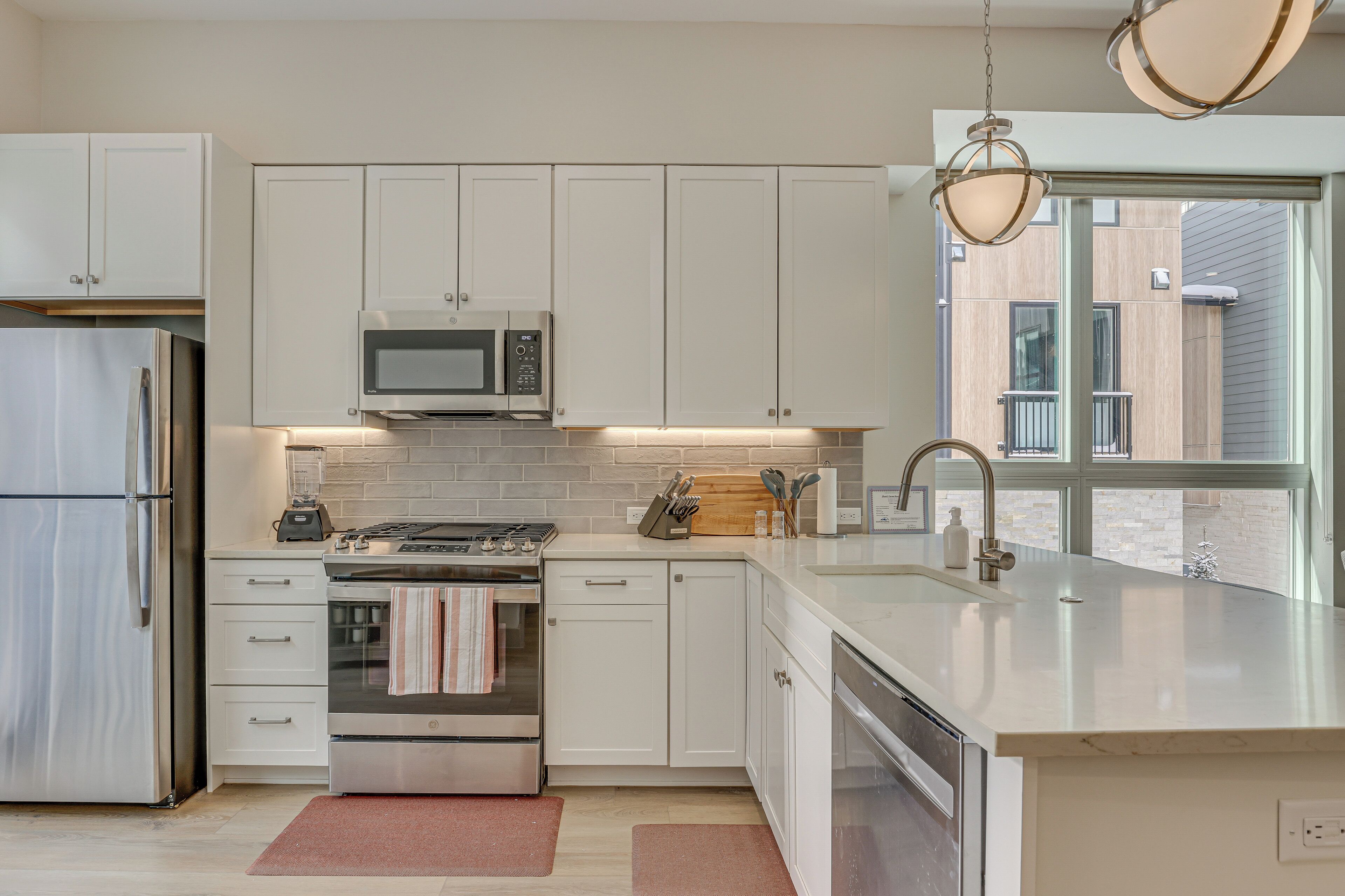 A modern kitchen with white cabinets, stainless steel appliances, and a large window providing a view of the building's exterior.