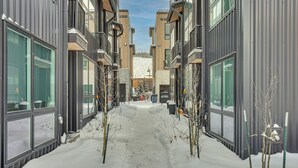A snow-covered pathway between modern buildings with large windows and a visible Christmas tree in the distance. - Silverthorne Townhome w/ Hot Tub: Walk to Town! (Silverthorne)