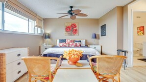 A bedroom with a bed, two wicker chairs, a table with a vase, a ceiling fan, and artwork on the walls. - Kailua-kona Condo Rental ~ 1/2 Mi to Golf Course! (Kailua-Kona)
