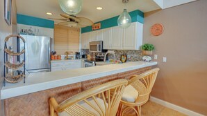 A kitchen with white cabinets, a stainless steel refrigerator, a microwave, and a sink. There are two wicker chairs at a bar counter. - Kailua-kona Condo Rental ~ 1/2 Mi to Golf Course! (Kailua-Kona)