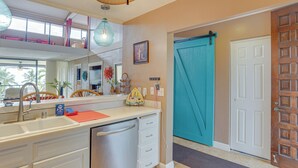 A kitchen with a dishwasher, a sink, and a countertop with a red mat. There is a large window with a view of palm trees. - Kailua-kona Condo Rental ~ 1/2 Mi to Golf Course! (Kailua-Kona)