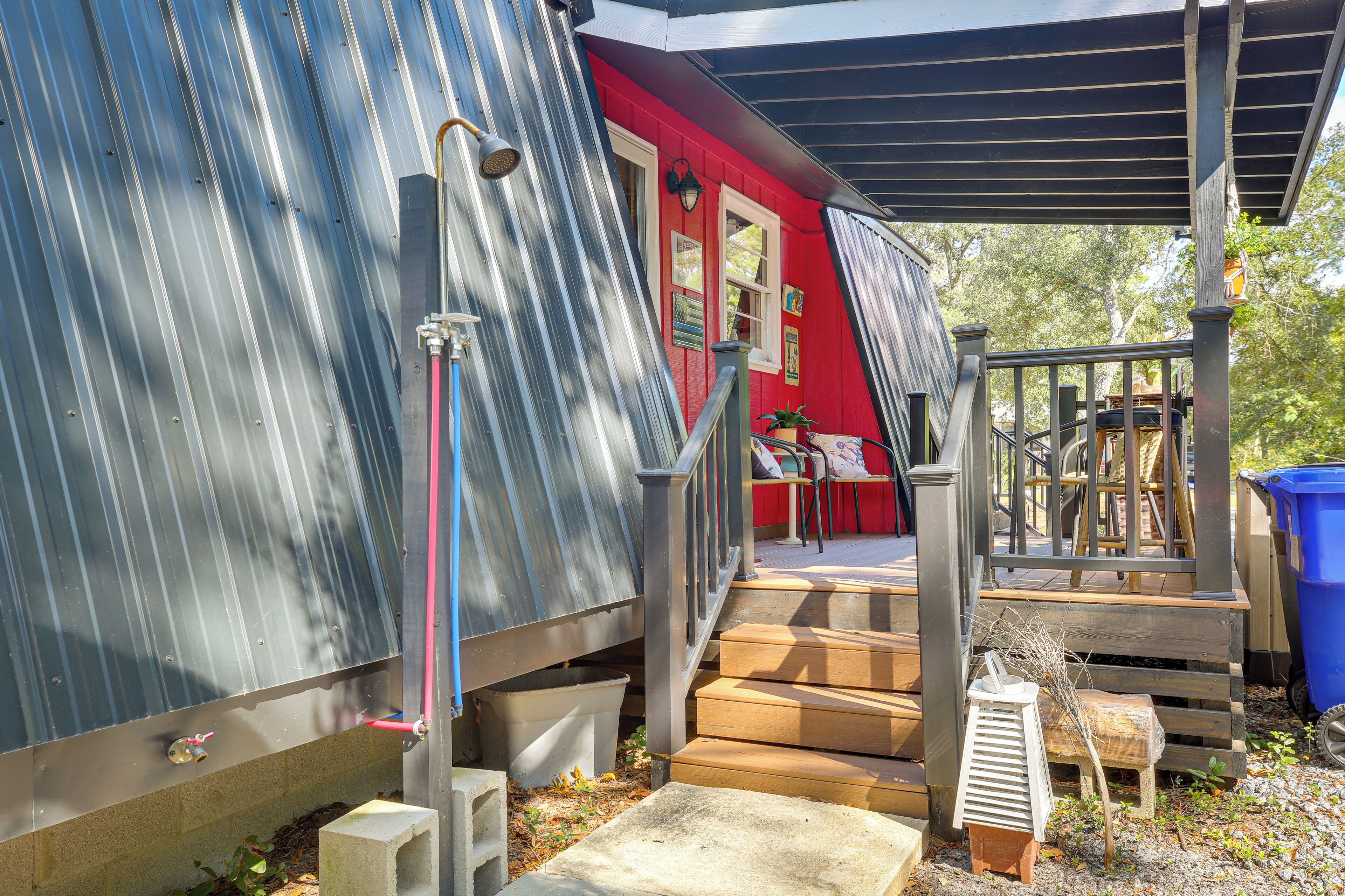 A metal structure with a red building in the background, wooden steps leading up to a covered area, and a blue trash bin.