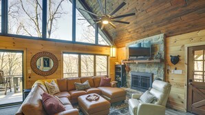 Ein gemütliches Blockhäuschen-Interieur mit einer Eckcouch, einem Kamin, einem Fernseher und einem großen Fenster mit Blick auf eine Terrasse.
