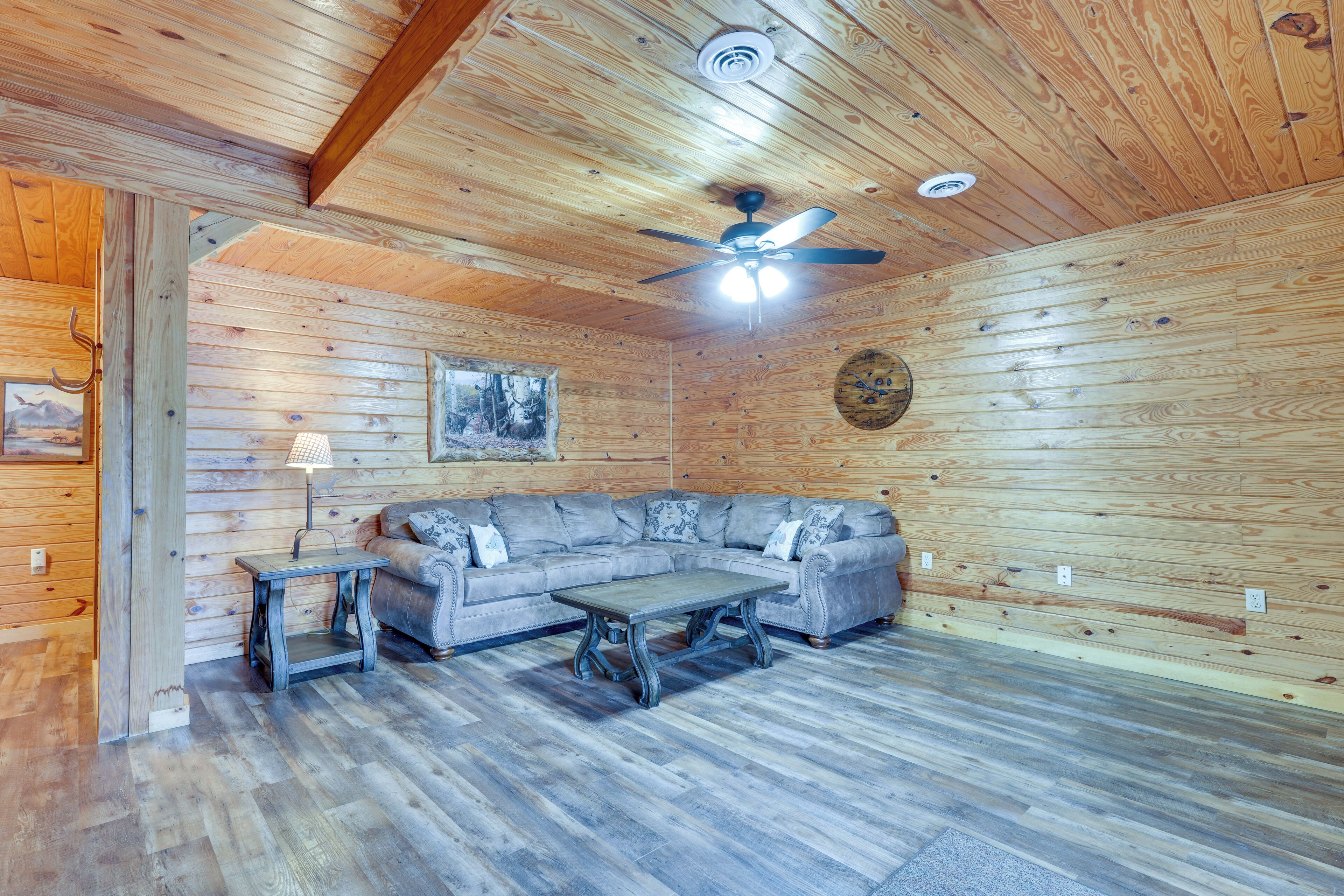 A log cabin interior with a wooden ceiling, a ceiling fan, a gray sectional sofa, a small table, and a lamp.