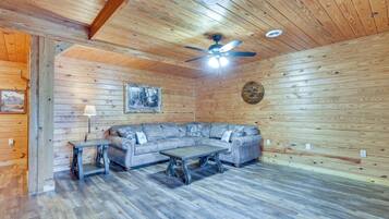 A log cabin interior with a wooden ceiling, a ceiling fan, a gray sectional sofa, a small table, and a lamp.