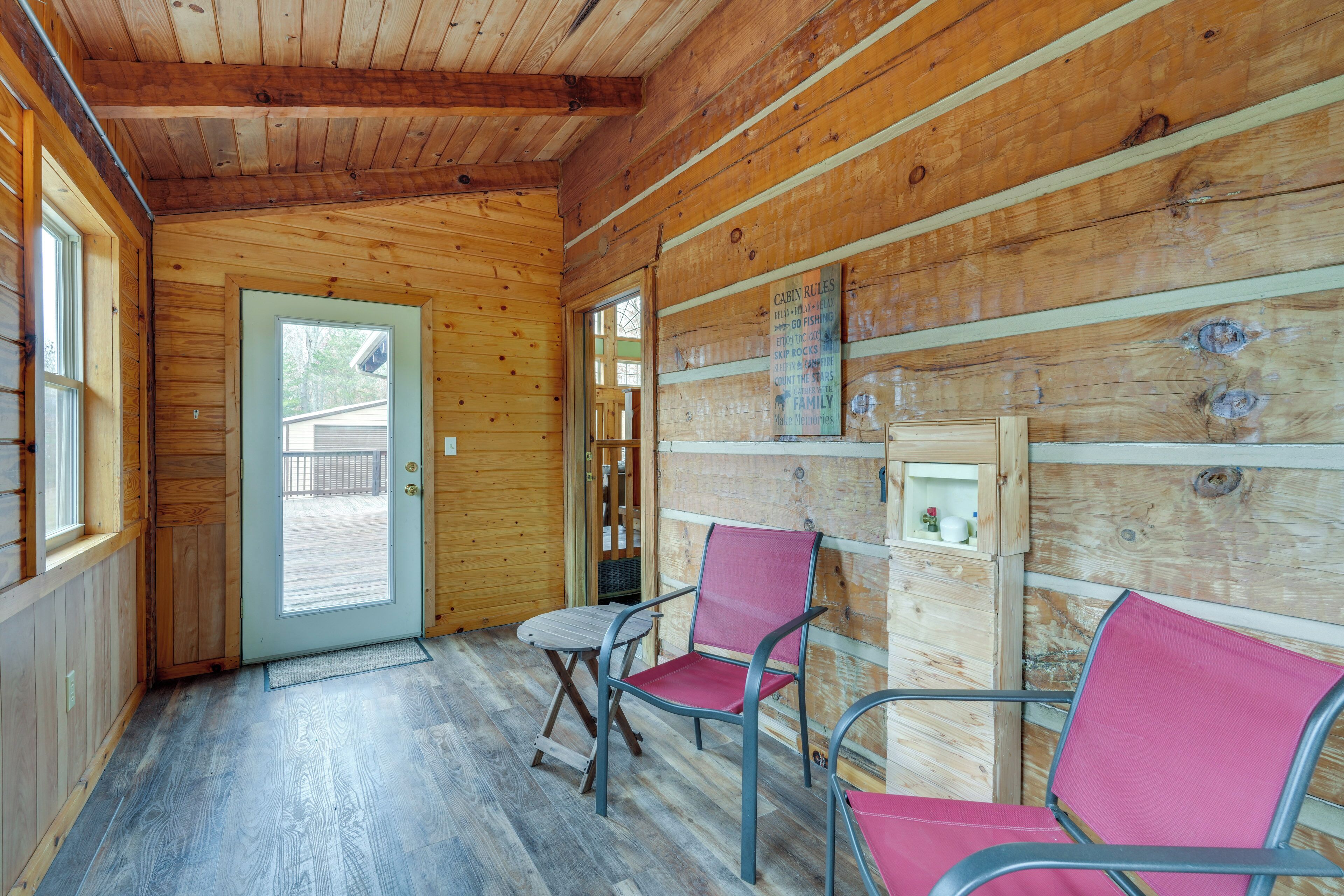 A cabin interior with wooden walls, a door leading outside, and two metal chairs with red cushions.