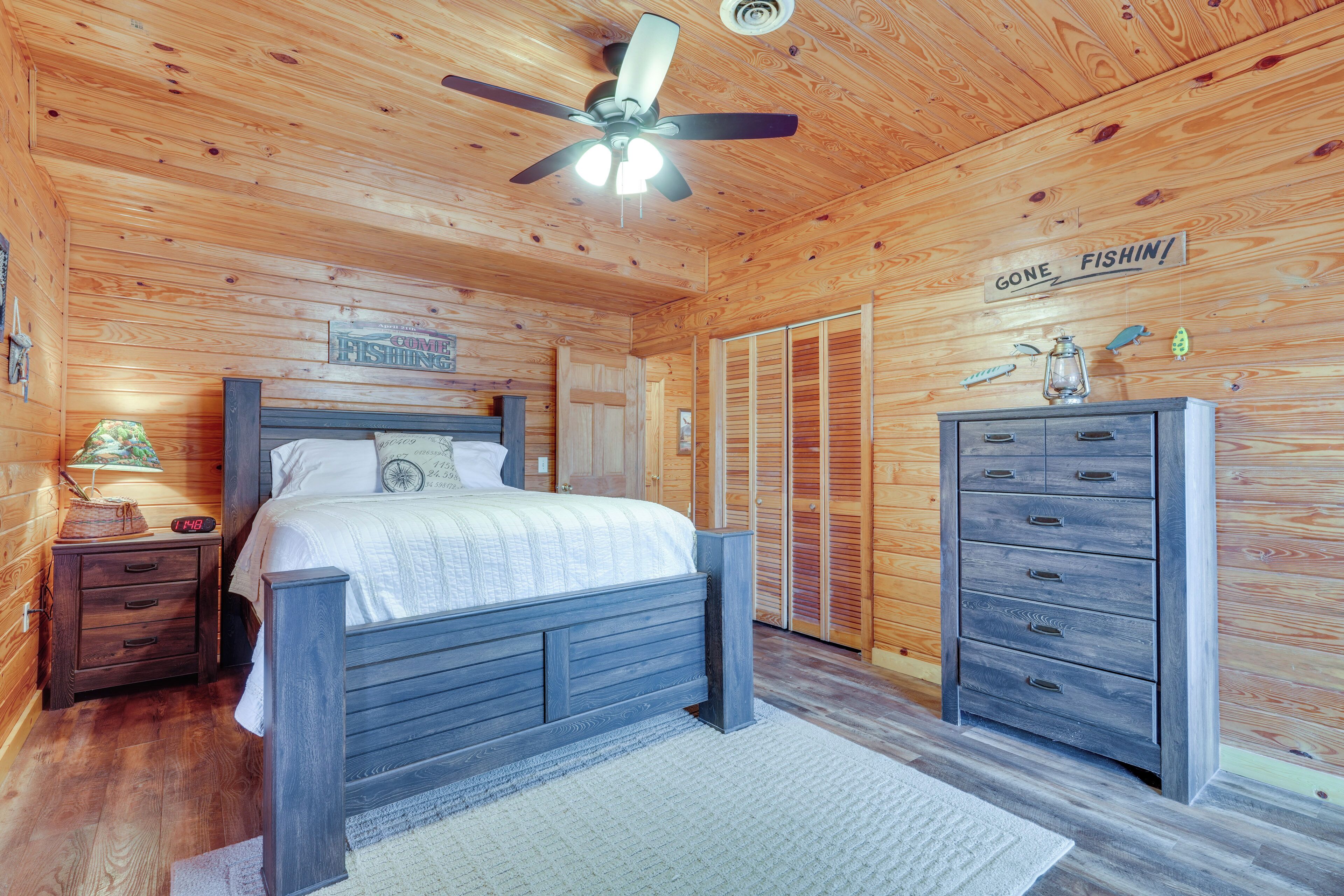 A wooden cabin bedroom with a bed, a dresser, a nightstand, a ceiling fan, and a sign on the wall.