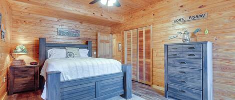 A wooden cabin bedroom with a bed, a dresser, a nightstand, a ceiling fan, and a sign on the wall.