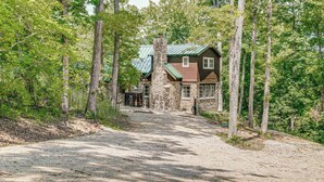 Un chalet rustique avec une cheminée en pierre, entouré de grands arbres et bordé d’une allée de gravier.