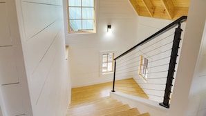 Un escalier avec des marches en bois et une rampe noire, menant à un deuxième étage aux murs blancs et au plafond en bois.