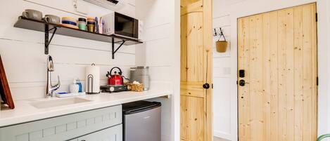 Een compacte keuken met witte kastjes, een ingebouwde magnetron en een compacte koelkast. Op de achtergrond is een houten deur en een groene tafel te zien.