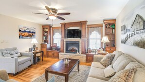 A living room with a fireplace, wooden bookshelves, a ceiling fan, and a large painting on the wall.