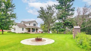 A house with a front porch, a circular stone fire pit, and a wooden shed in a grassy yard.