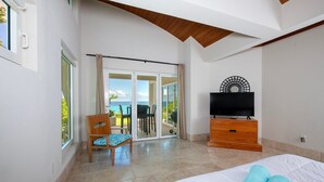 A hotel room with a bed, a TV, a wooden dresser, a chair, and a sliding glass door leading to a balcony with a view of the ocean. - Sky Beach Club Villa Waterfall 2 4 Bedroom Home (Governor's Harbour)