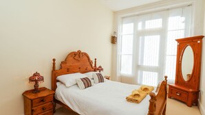 Une chambre à coucher avec un lit en bois, une table de chevet, une commode et une grande fenêtre ornée de rideaux légers.