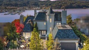 A large, multi-story house with a prominent turret, situated by a lake with a marina in the background. - Tudor Revival Castle with Bay Views 5bd / 5 bath (Mill Valley)