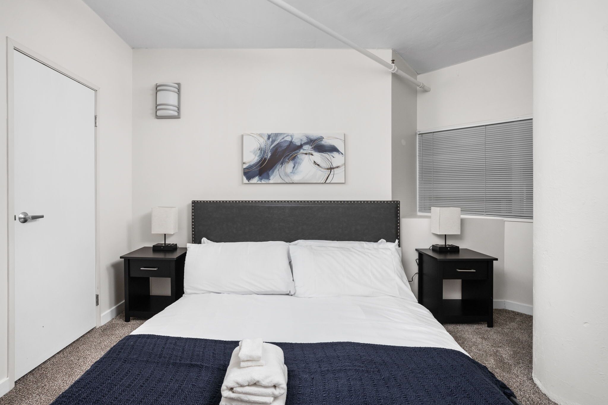 A bedroom with a bed, two nightstands, a wall-mounted lamp, a window with blinds, and a painting on the wall.